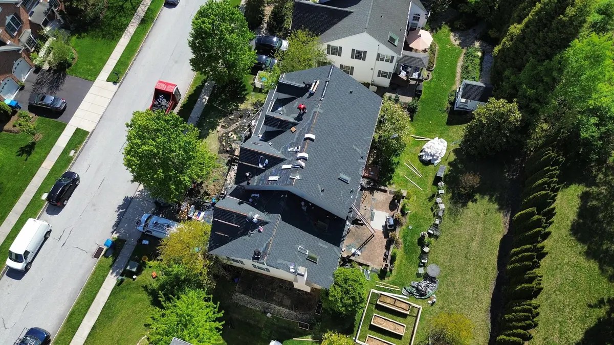 Aerial view of a large home with new dark shingles across most of the roof, crew working and materials still stacked on the slopes