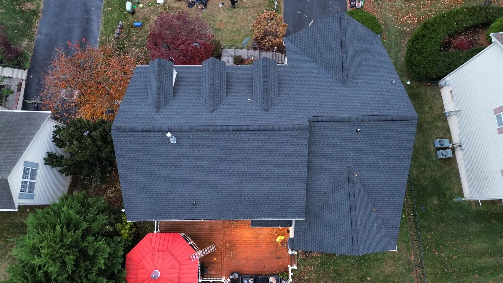 Top down autumn aerial of a completed blue gray shingle roof with a red patio umbrella and wooden deck below