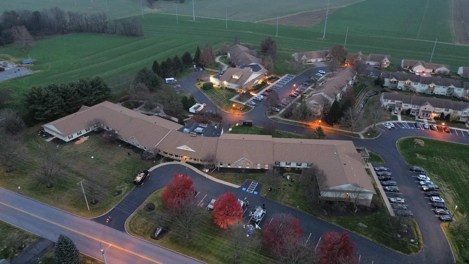 Dusk aerial of a long multi-family building with a finished tan shingle roof, lit windows and a parking lot lined with red trees
