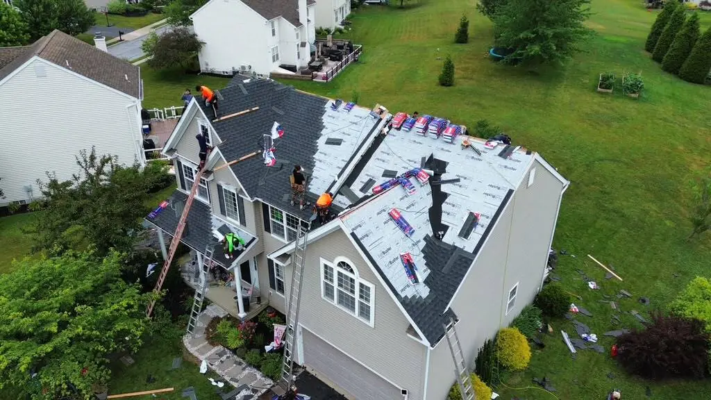 Aerial view of five roofers installing dark shingles over white underlayment on a tan siding home with ladders up at the eaves