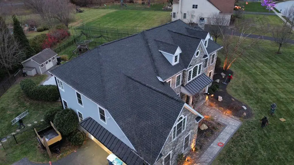 Evening aerial view of a stone and siding home with a finished dark shingle roof, a standing seam porch roof and landscape lighting across the front beds