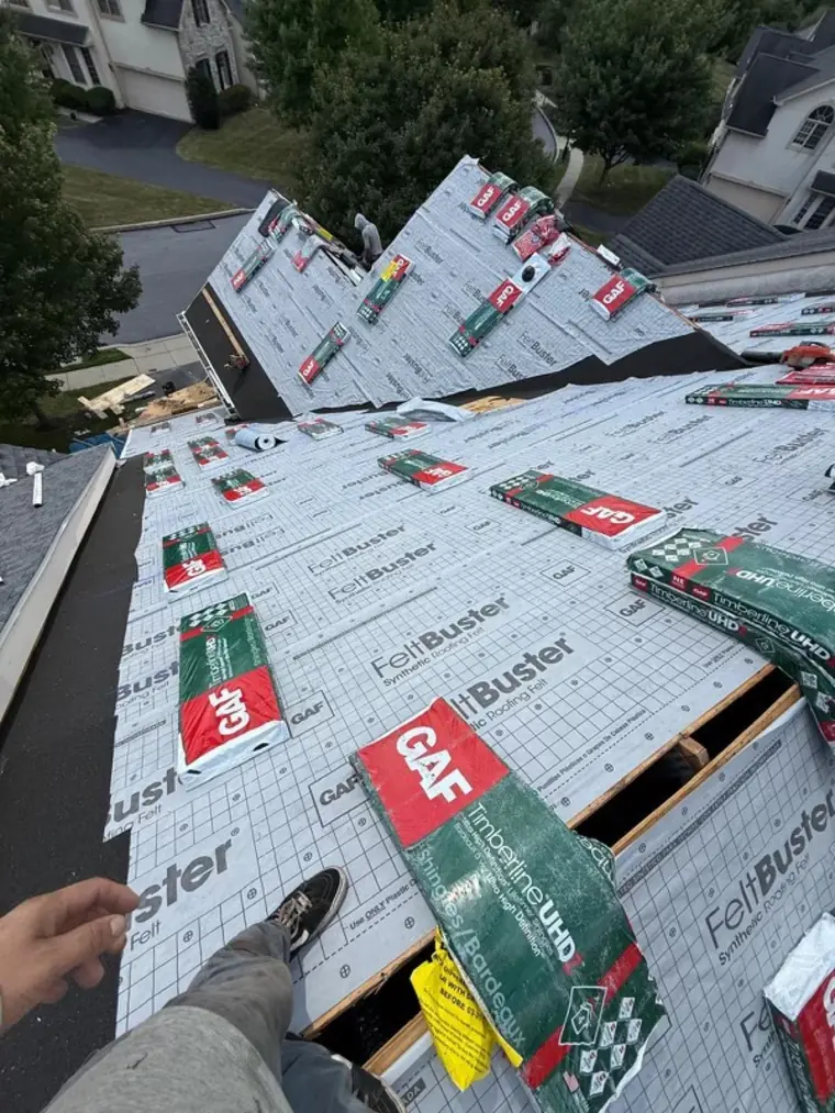 View from a roofer standing on a roof papered in GAF FeltBuster underlayment, with GAF Timberline shingle bundles staged around him and the neighborhood below