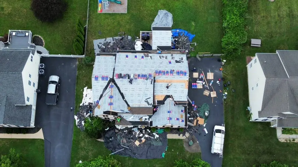 Top down aerial of a roof stripped to the deck and covered in underlayment, with tarps and debris staged on the driveway below
