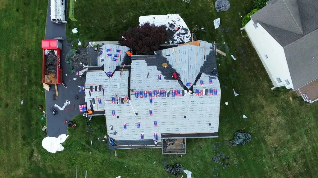 Top down aerial of a house stripped to fresh underlayment with shingle bundles spread across every slope and a red dump trailer in the driveway