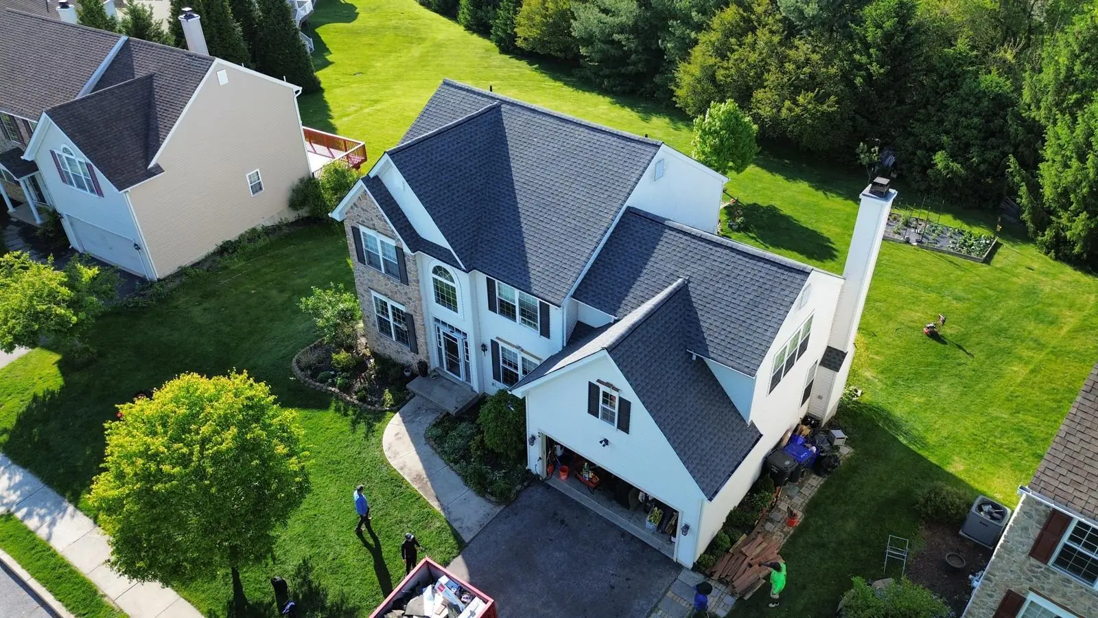 Sunny aerial view of a stone and white siding home with a completed dark shingle roof and the crew packing up in the driveway