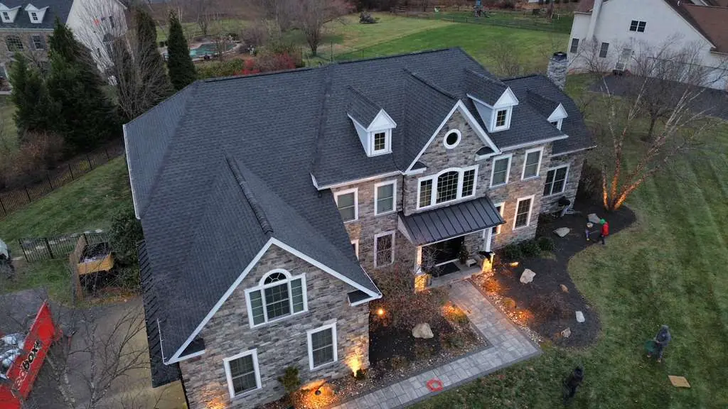 Evening aerial of a large stone home with a finished dark shingle roof, three dormers and landscape lighting along the walkway