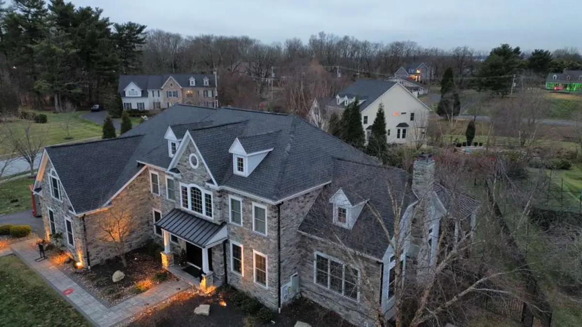 Evening aerial view of a large stone home with a finished dark shingle roof, dormers and a standing seam metal porch roof