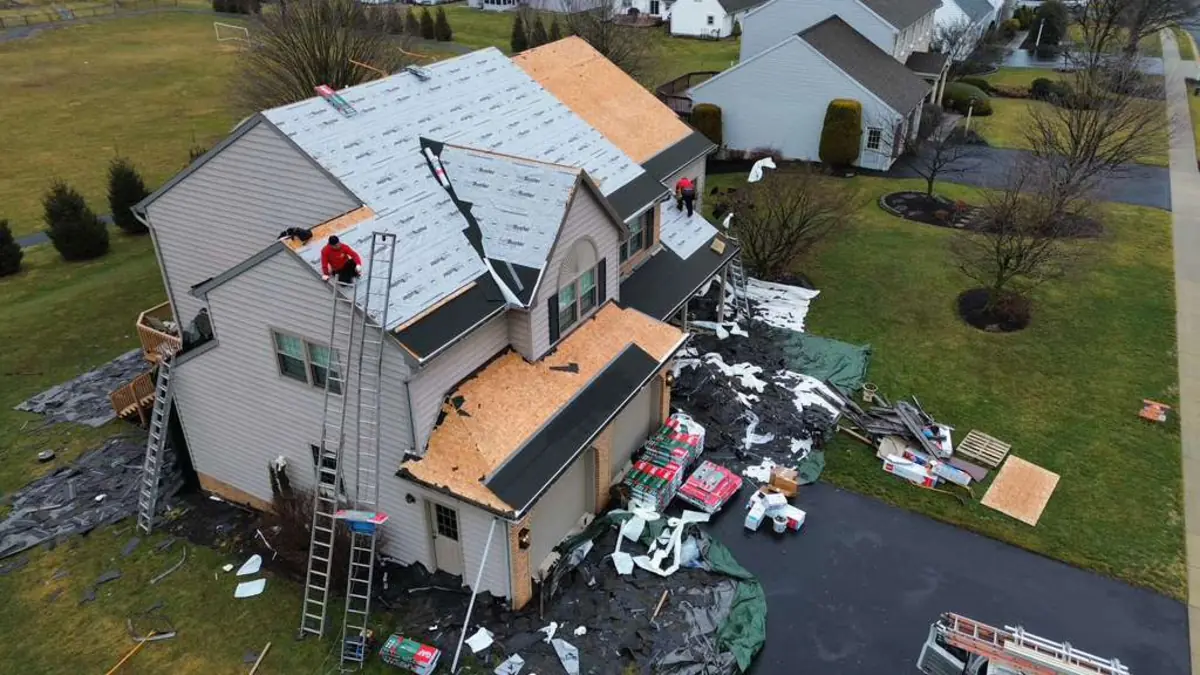 Aerial view of a home mid tear off with bare roof decking exposed, crew on ladders and tarps laid over the lawn and driveway to catch debris