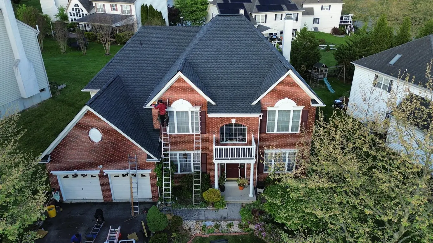 Roofer in a red shirt on an extension ladder at the gutter line of a red brick home with a new charcoal shingle roof above